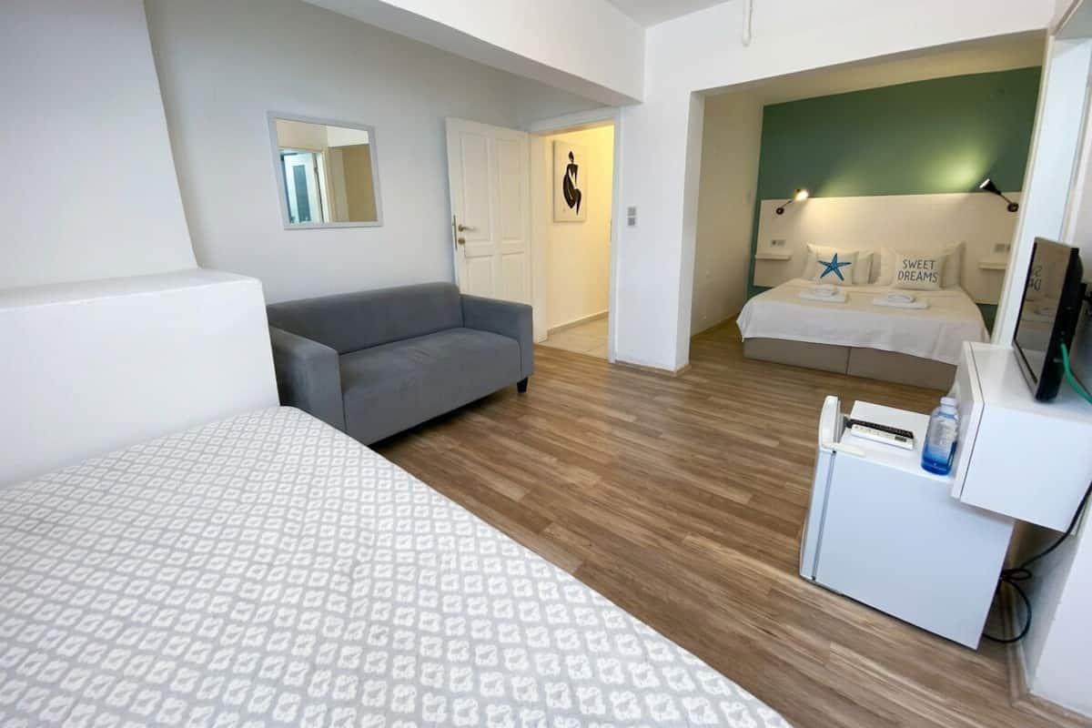 Ein modernes Hotelzimmer mit einem großen Bett, einem grauen Sofa, einem kleinen Schreibtisch mit einem Computer und einem separaten Schlafbereich mit Bett und Nachttisch.