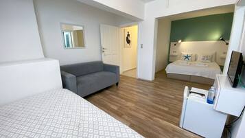 Una habitación de hotel moderna con una cama grande, un sofá gris, un escritorio pequeño con una computadora y una zona de descanso separada con cama y mesita de noche.