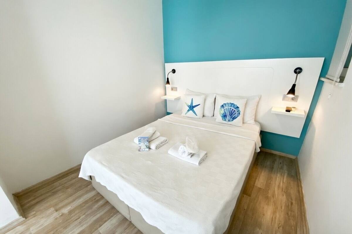 Ein ordentlich bezogenes Bett mit weißen Leinen und einem Kopfkissen mit Stern und Muschel, ein Nachttisch mit Lampe und eine wandfarbige Akzentwand in Türkis.