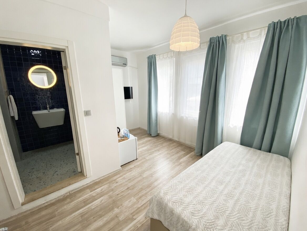 Quarto moderno com cama, mesa de cabeceira, luminária pendente e vista para um banheiro com pia e espelho.