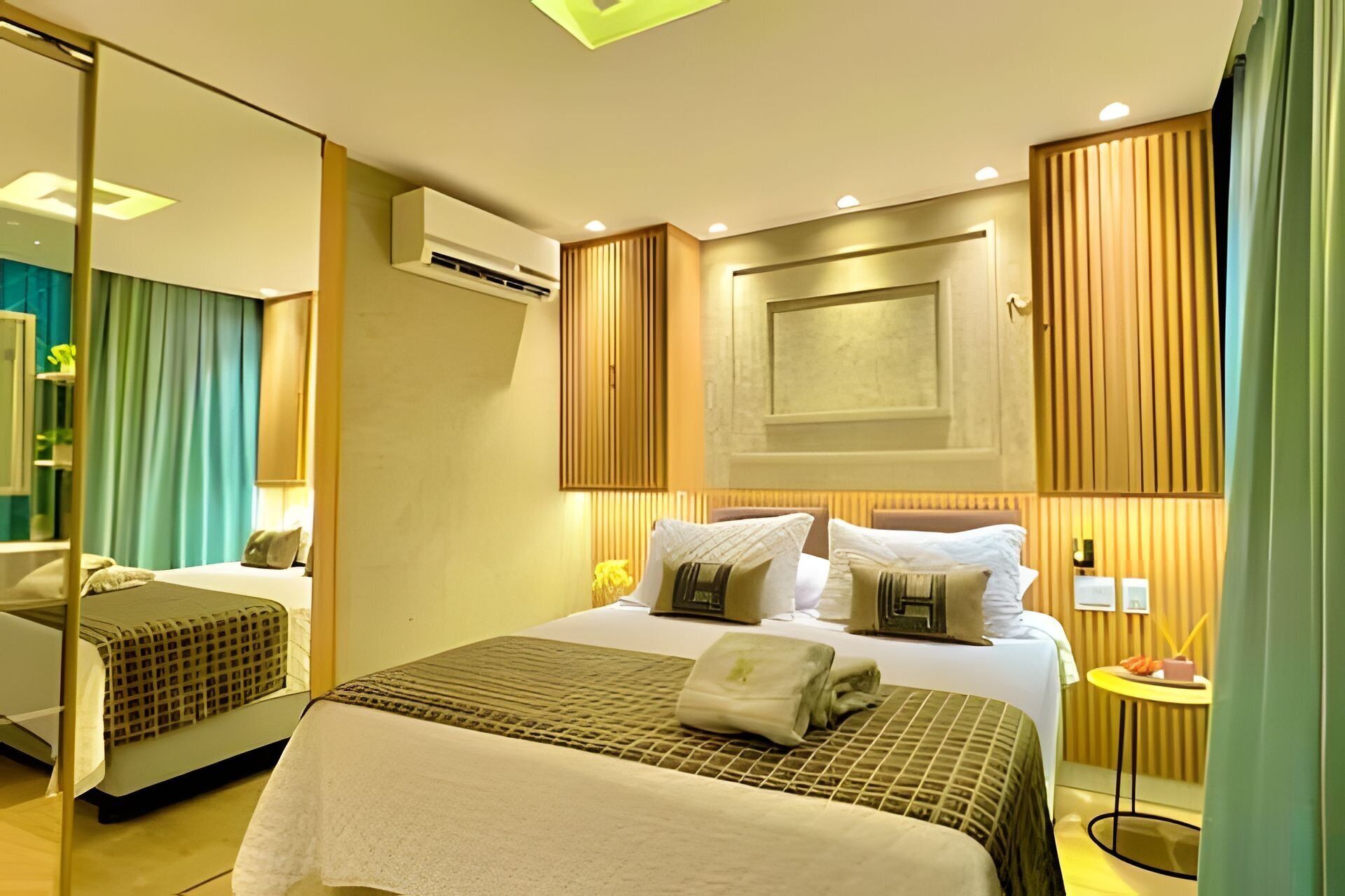 A hotel room with a large bed, a smaller bed, a bedside table with a lamp, an air conditioning unit, and a mirror.