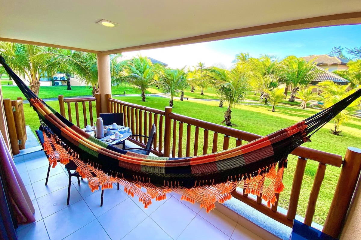 Un balcon avec un hamac, une table et des chaises, et une vue sur une pelouse bien entretenue et des palmiers.