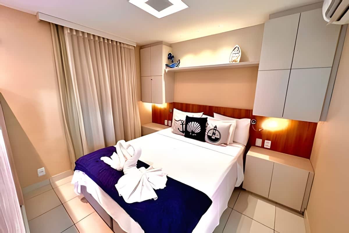A hotel room with a bed, curtains, a shelf with shoes, and a headboard with built-in lighting.