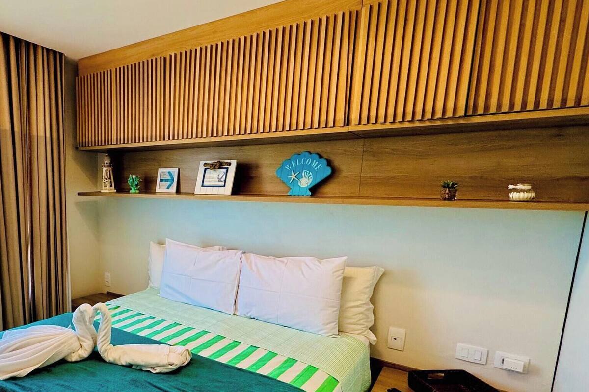 Une chambre d’hôtel avec un lit, une fenêtre avec des rideaux, une tête de lit en bois et une étagère contenant divers objets.