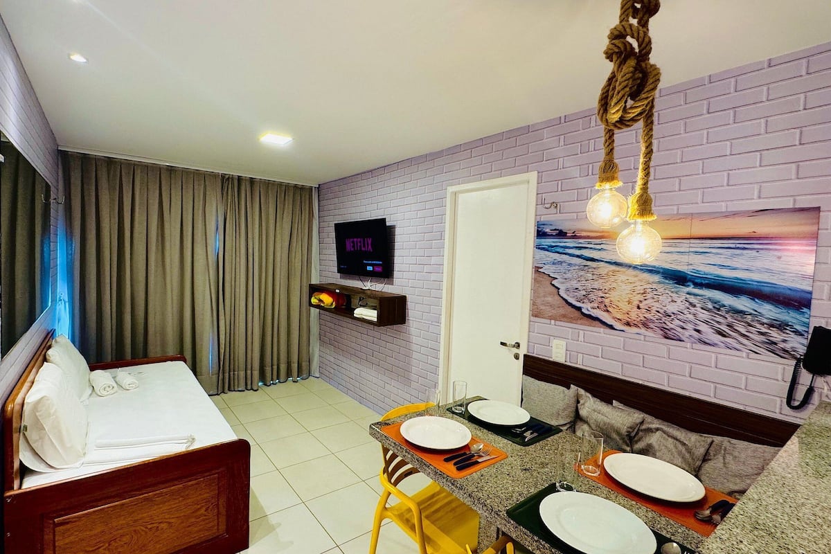 Une chambre d’hôtel comprenant un lit, un téléviseur fixé au mur, une table à manger avec des chaises et un tableau représentant une scène de plage.