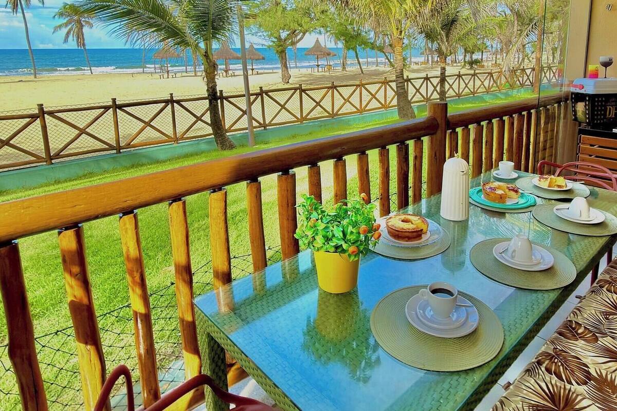 Un balcon avec une table dressée pour deux personnes, donnant sur une plage bordée d’une clôture et de palmiers.