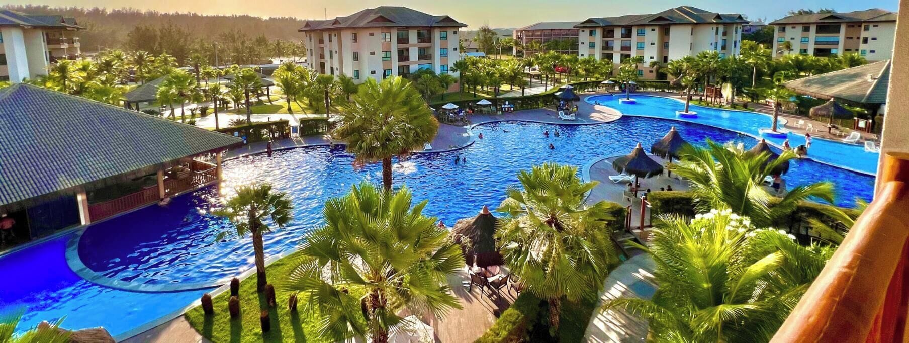 Un resort con una amplia piscina central, palmeras y varias edificaciones de hotel.