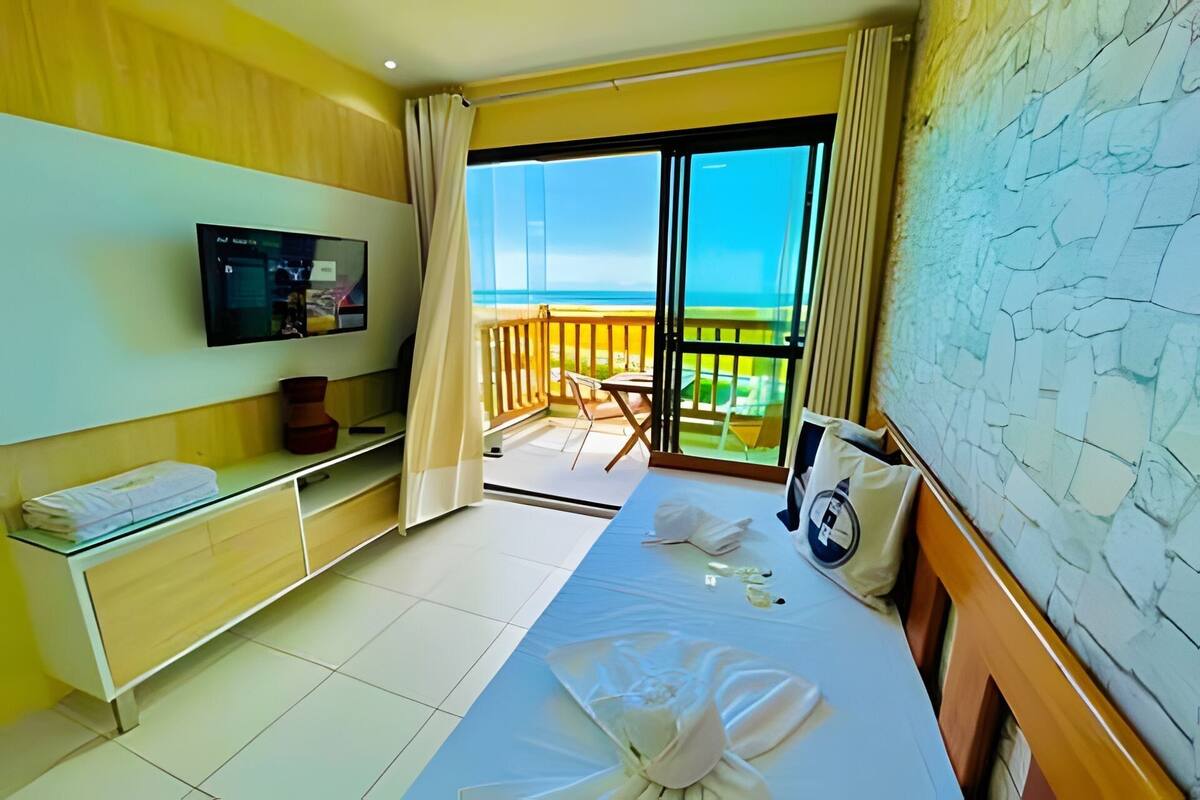 Une chambre d’hôtel avec un lit, une télévision, un balcon donnant sur la mer et une petite table sur laquelle est posé un vase.