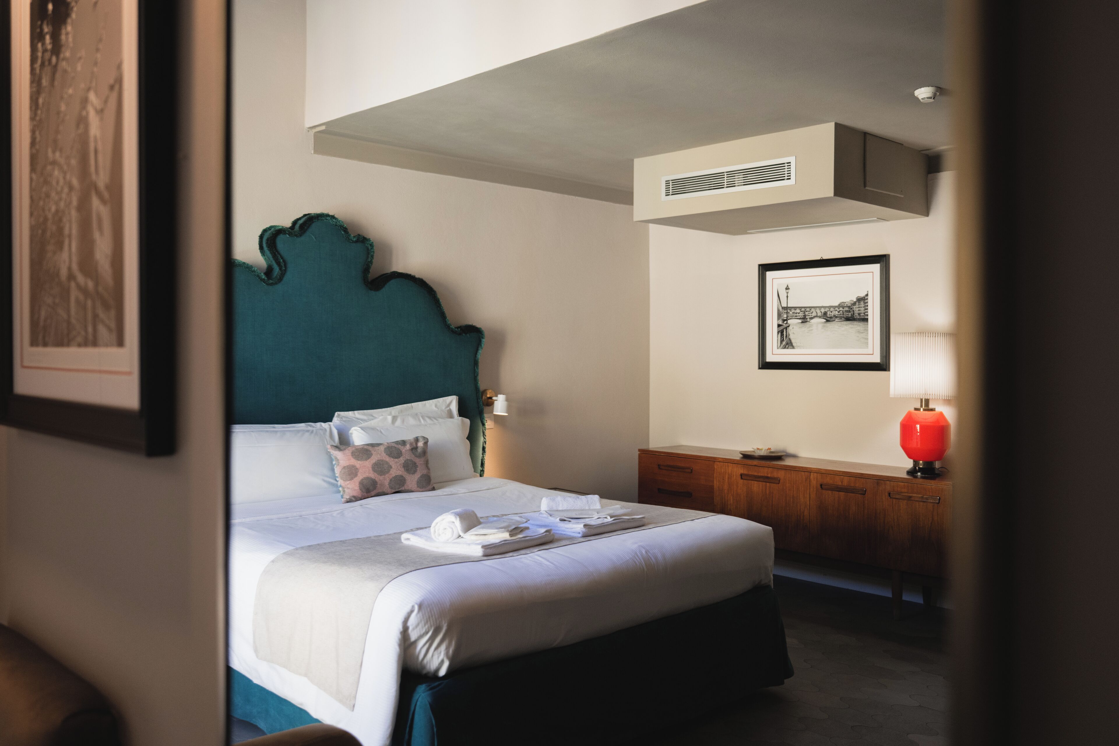 Una cama bien hecha con almohadas y una toalla enrollada, una mesita de noche de madera con una lámpara, y un cuadro en la pared.
