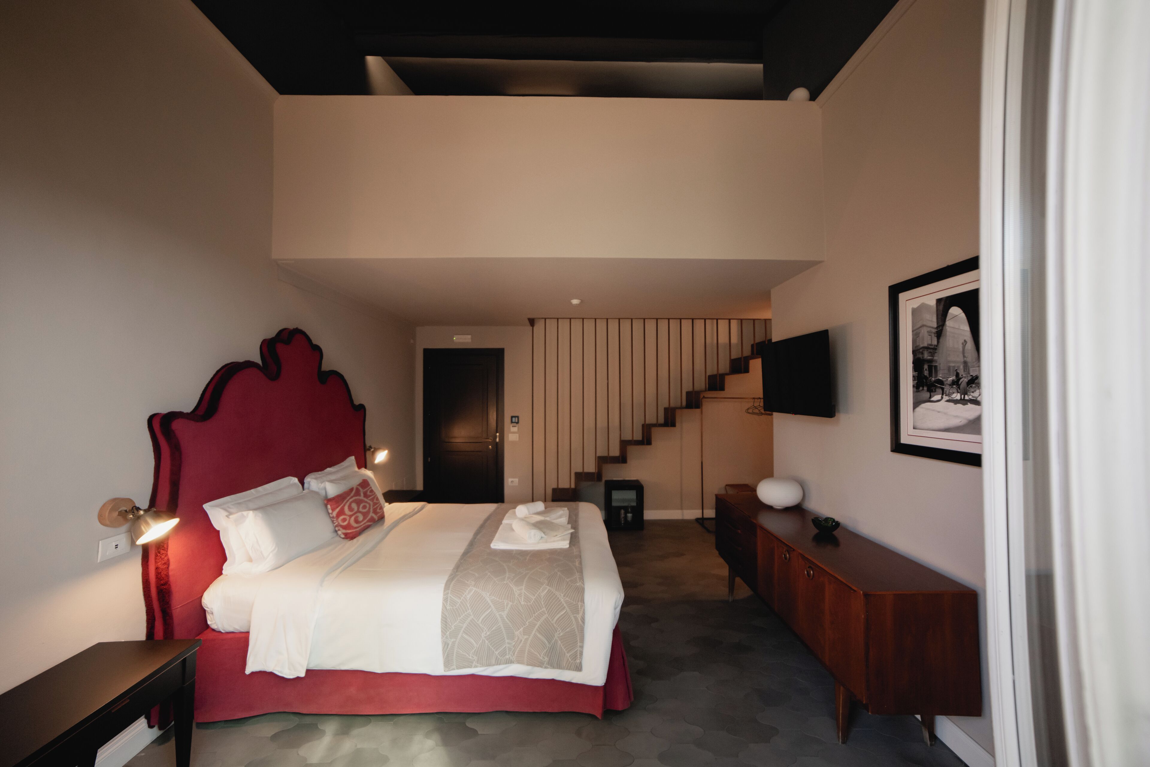 A modern bedroom with a red headboard, a bed with white linens, a wooden nightstand, a bedside table, a wooden dresser, and a flat-screen TV mounted on the wall.