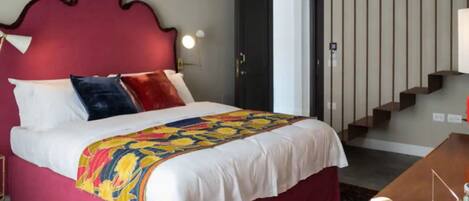 A hotel room with a large bed, a red headboard, a wooden desk, and a staircase in the background.