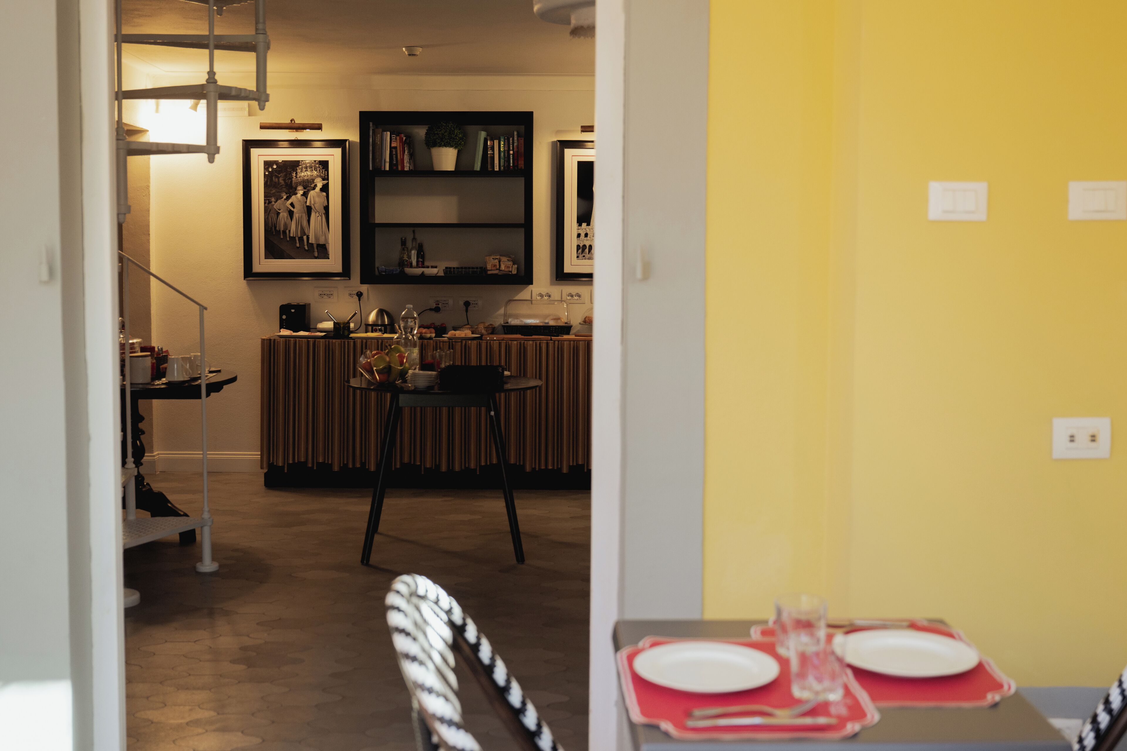 A dining area with a table set for two, a kitchen in the background, and a staircase leading to another level.
