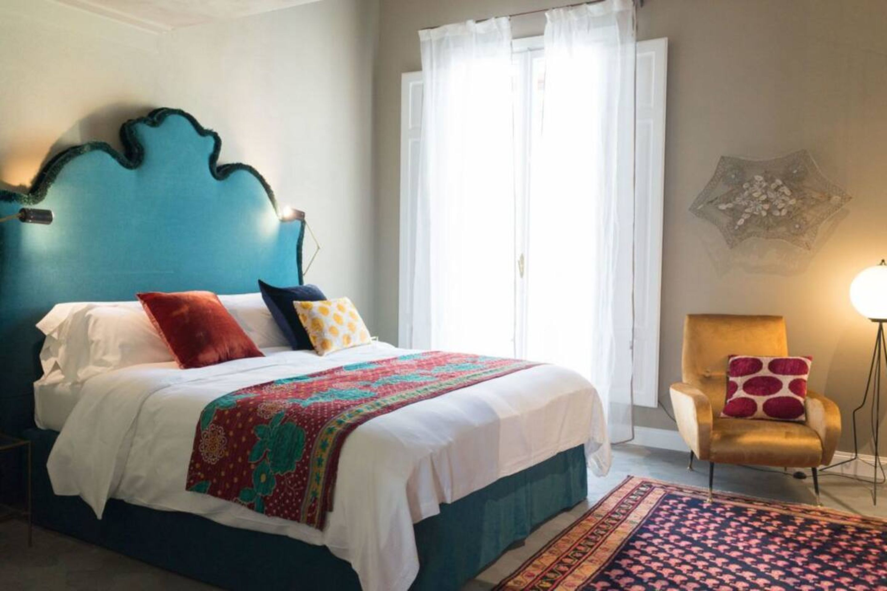 A bedroom with a teal headboard, a bed with white bedding and colorful pillows, a gold armchair, and a patterned rug.