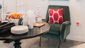 Una sala de estar moderna con un sillón verde, una mesa redonda con platos y frutas, una escalera en espiral blanca y una pintura de una planta tropical grande en la pared.