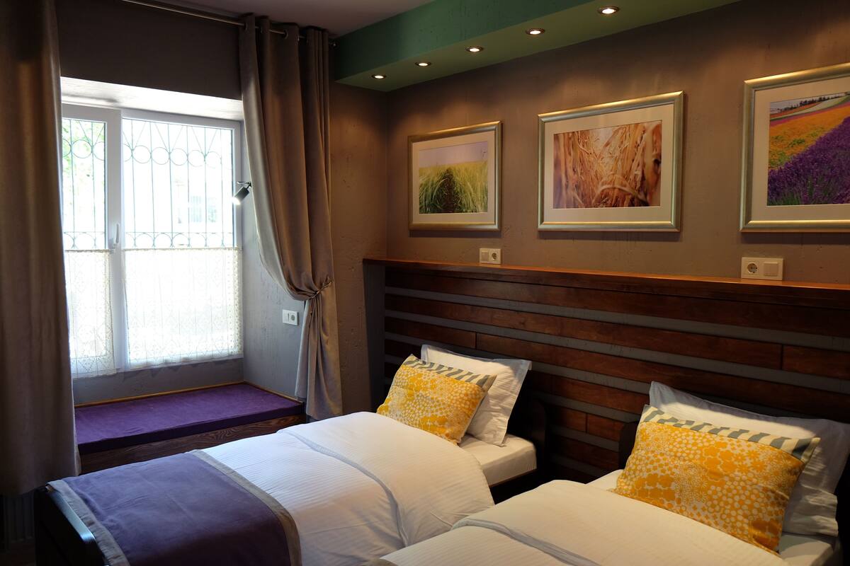 A room with two beds, a window with curtains, and three framed pictures on the wall.