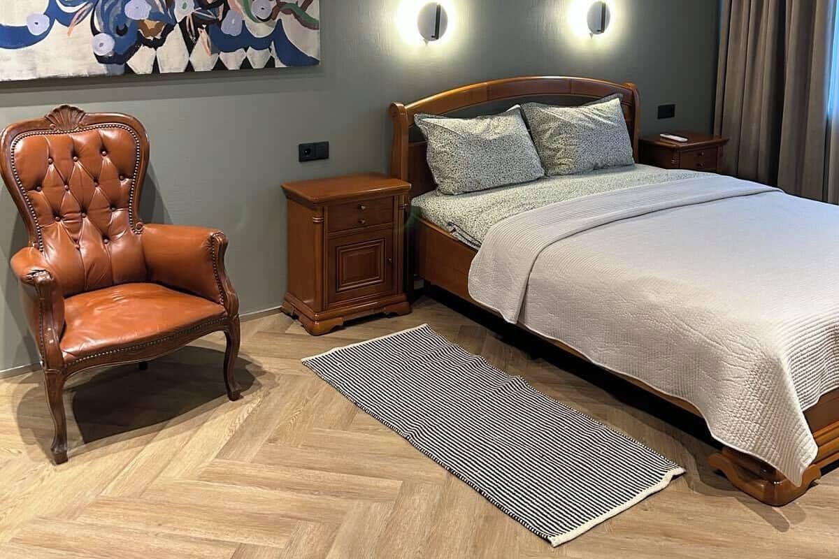 A bedroom with a bed, a chair, a nightstand, a wall-mounted light, and a painting.