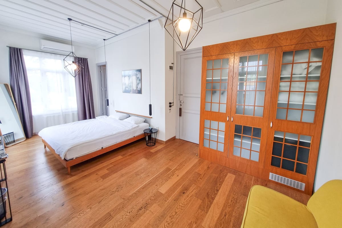 A bedroom with a wooden floor, a bed, a desk, a chair, a wardrobe, and a hanging light fixture.