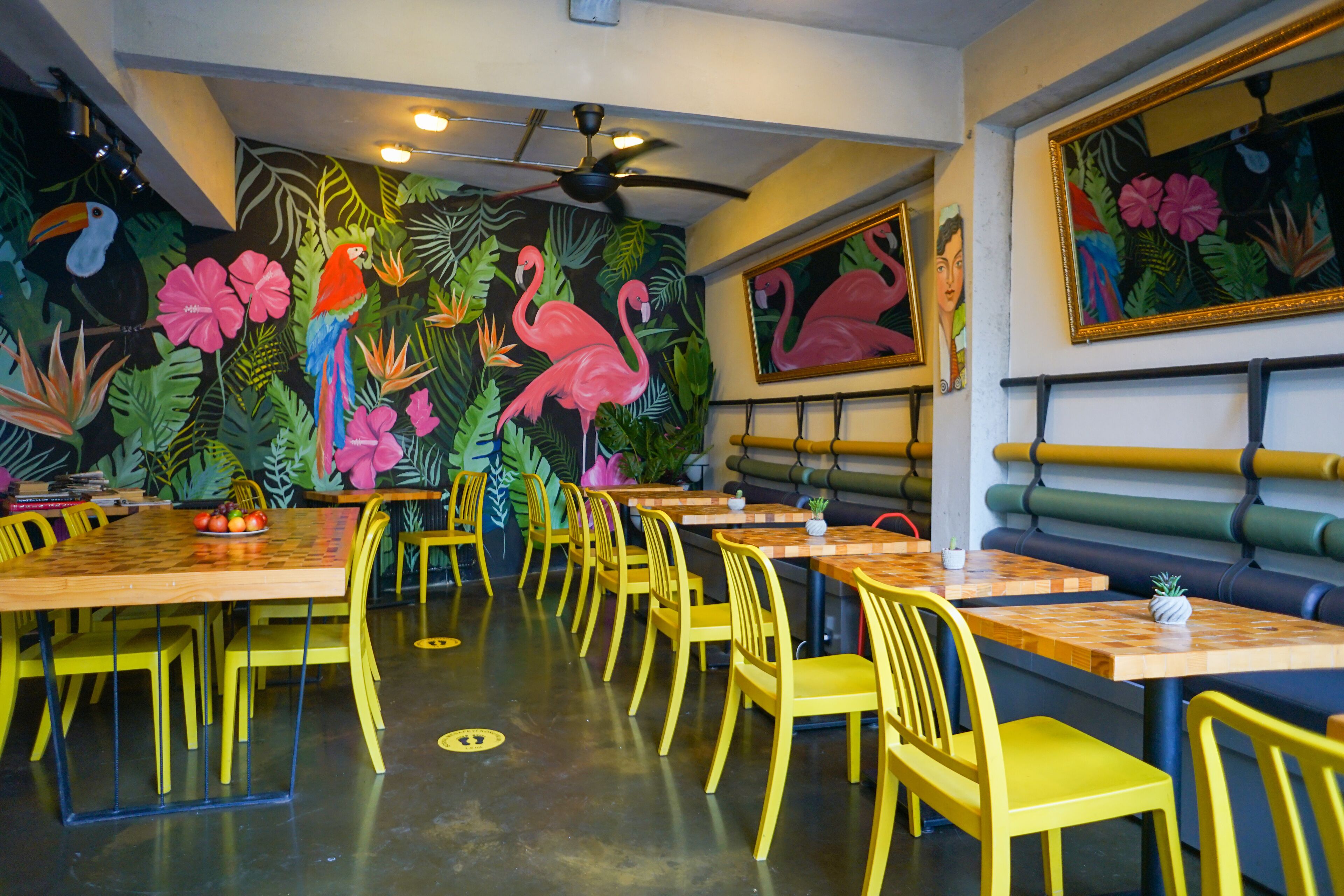 A restaurant with yellow chairs and tables, a ceiling fan, and a mural with tropical plants and flamingos.