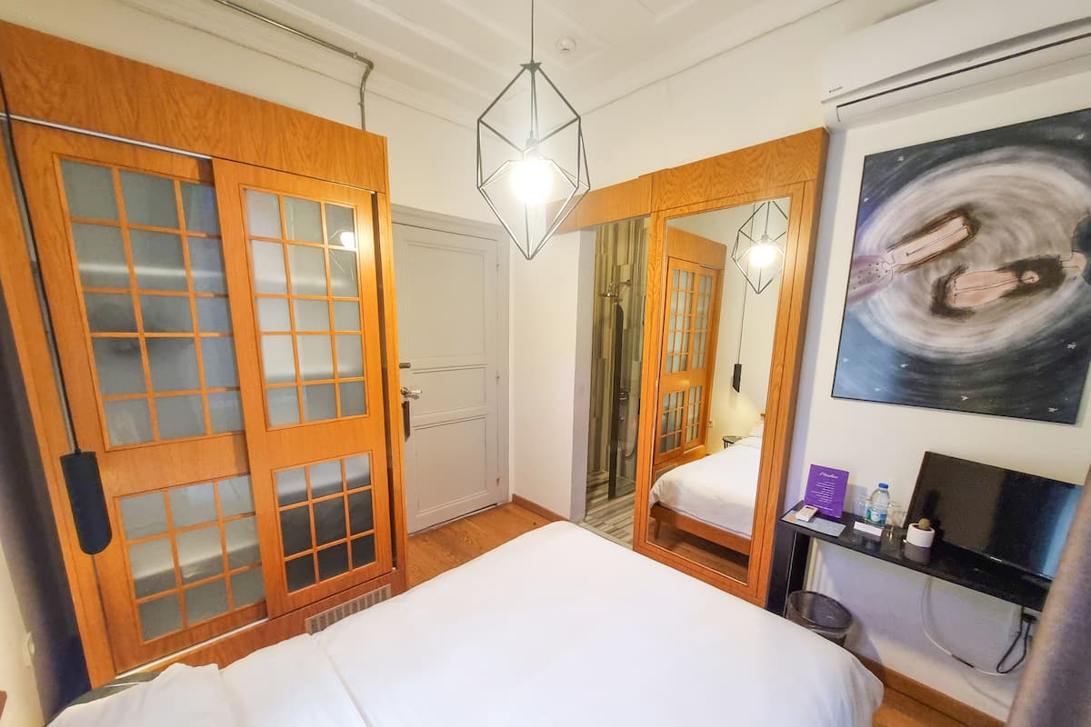 A hotel room with a large bed, wooden wardrobe with glass doors, a shower, a bedside table with a lamp, and a painting on the wall.