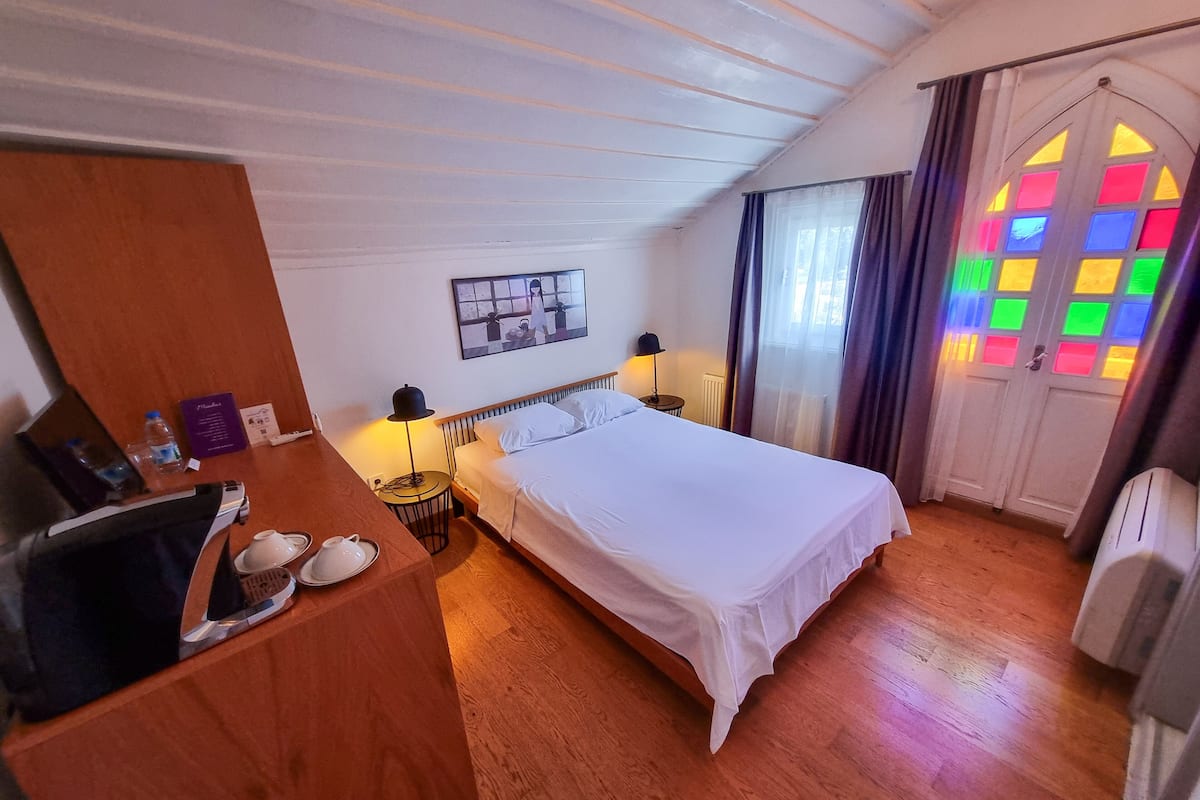 A hotel room with a wooden floor, a bed with white linens, a wooden wardrobe, a small table with a lamp, and a window with colorful stained glass.