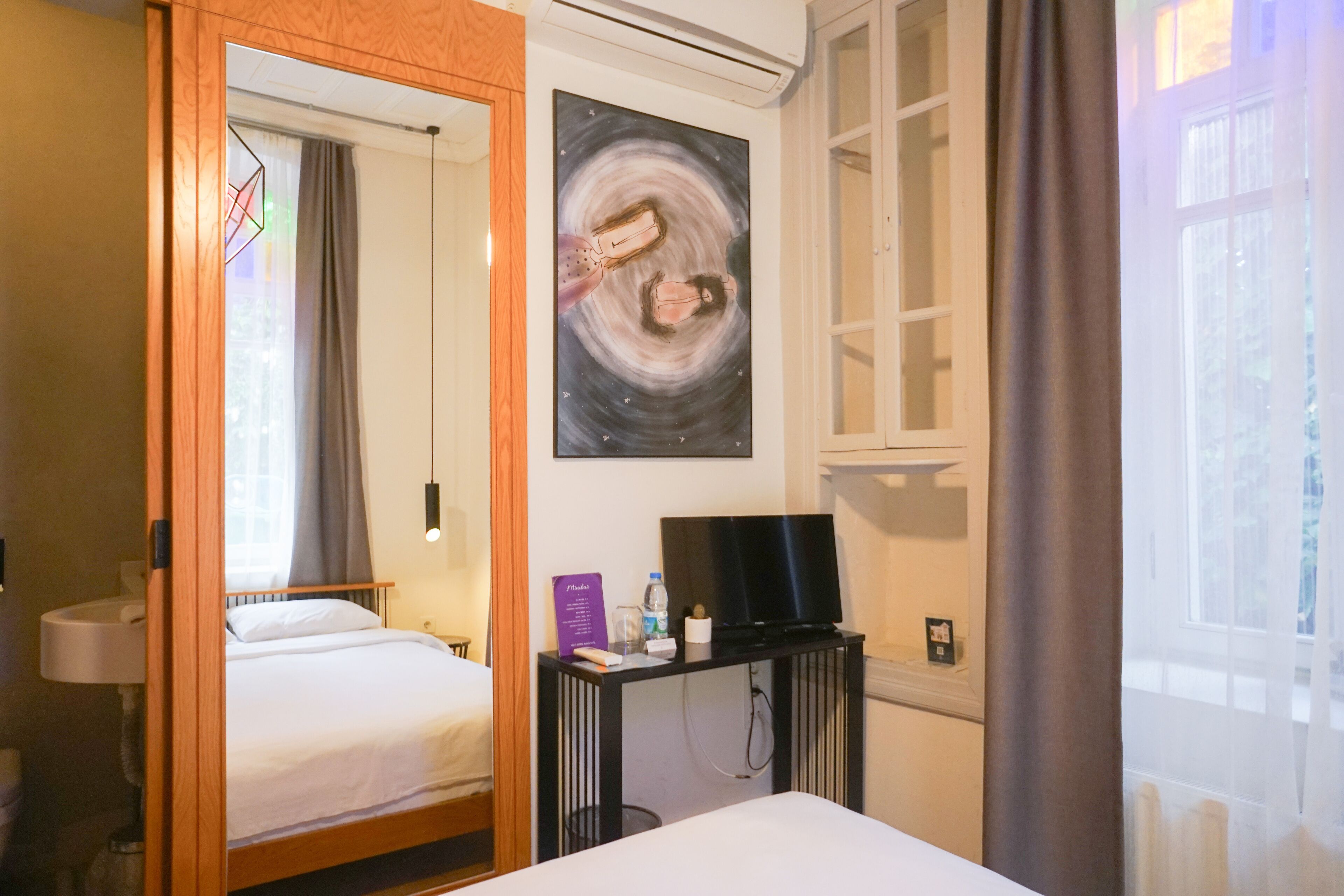 A hotel room with a bed, a desk with a television, a painting on the wall, and a window with curtains.
