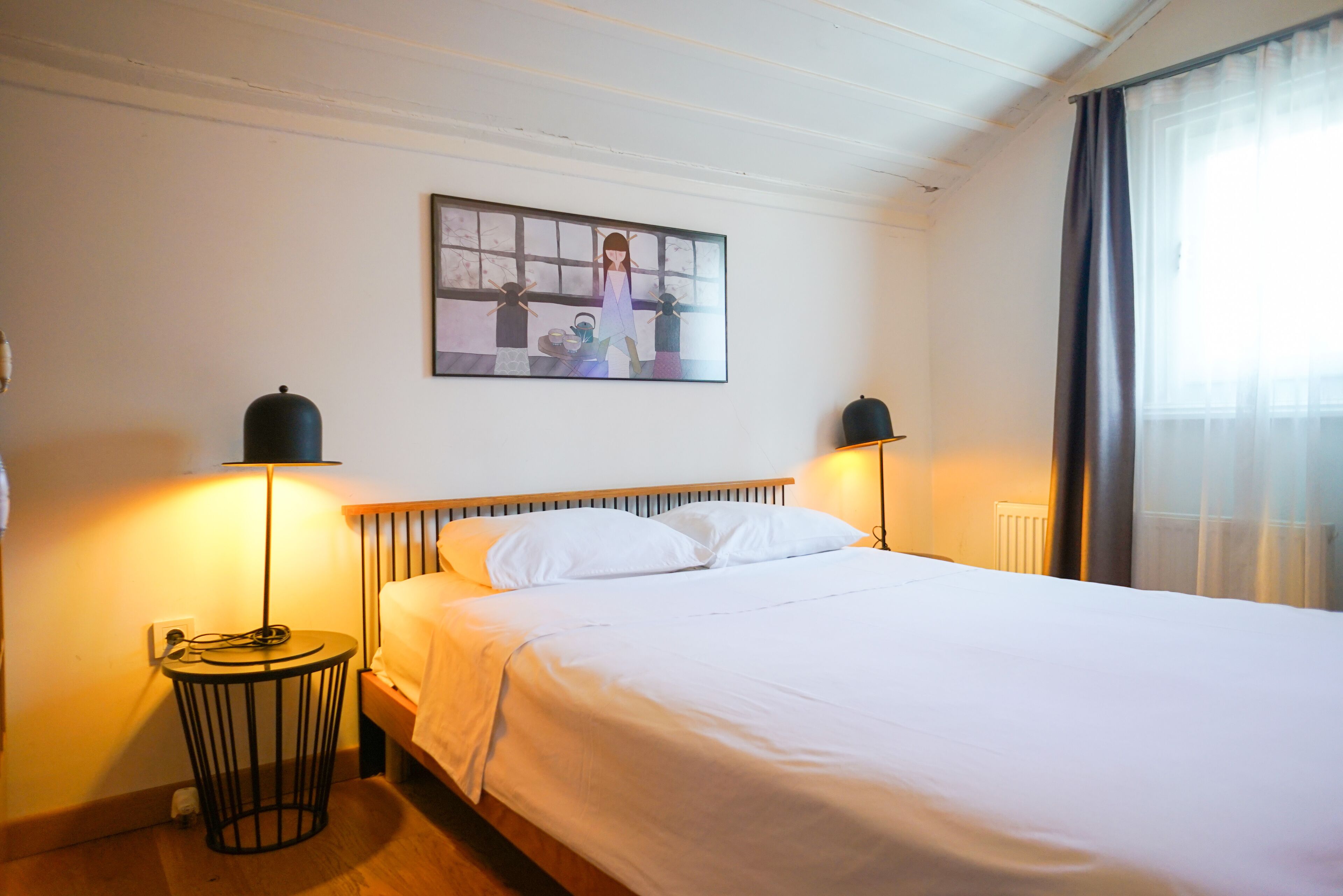 A neatly made bed with white linens, two bedside lamps, a framed artwork above the bed, and a window with curtains.