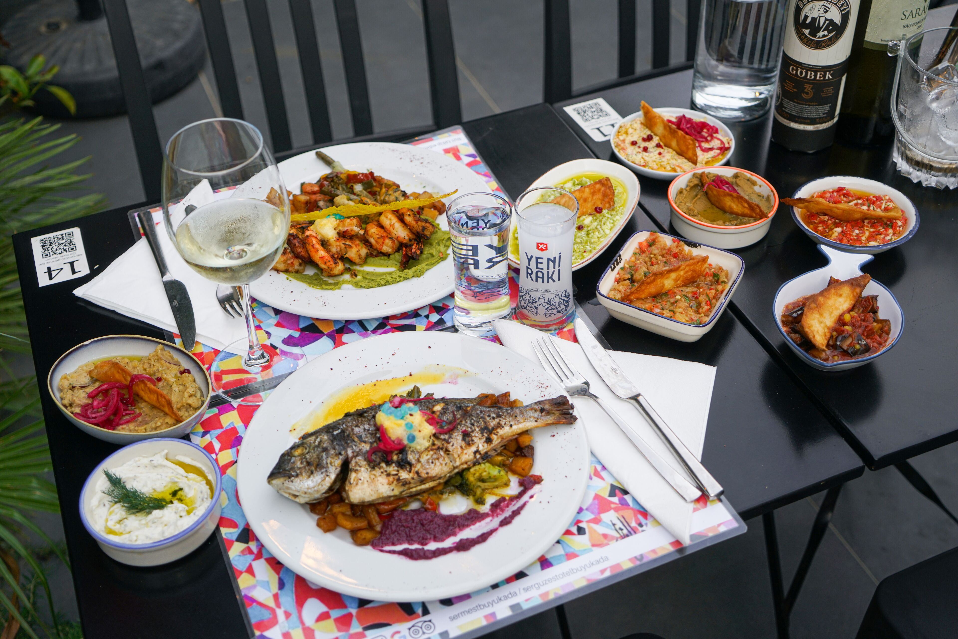A table set with various dishes, including a whole fish, bowls of dips, and a bottle of wine.