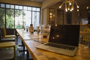 Una computadora portátil sobre una mesa de madera en una cafetería con ventanas amplias y una lámpara colgante.