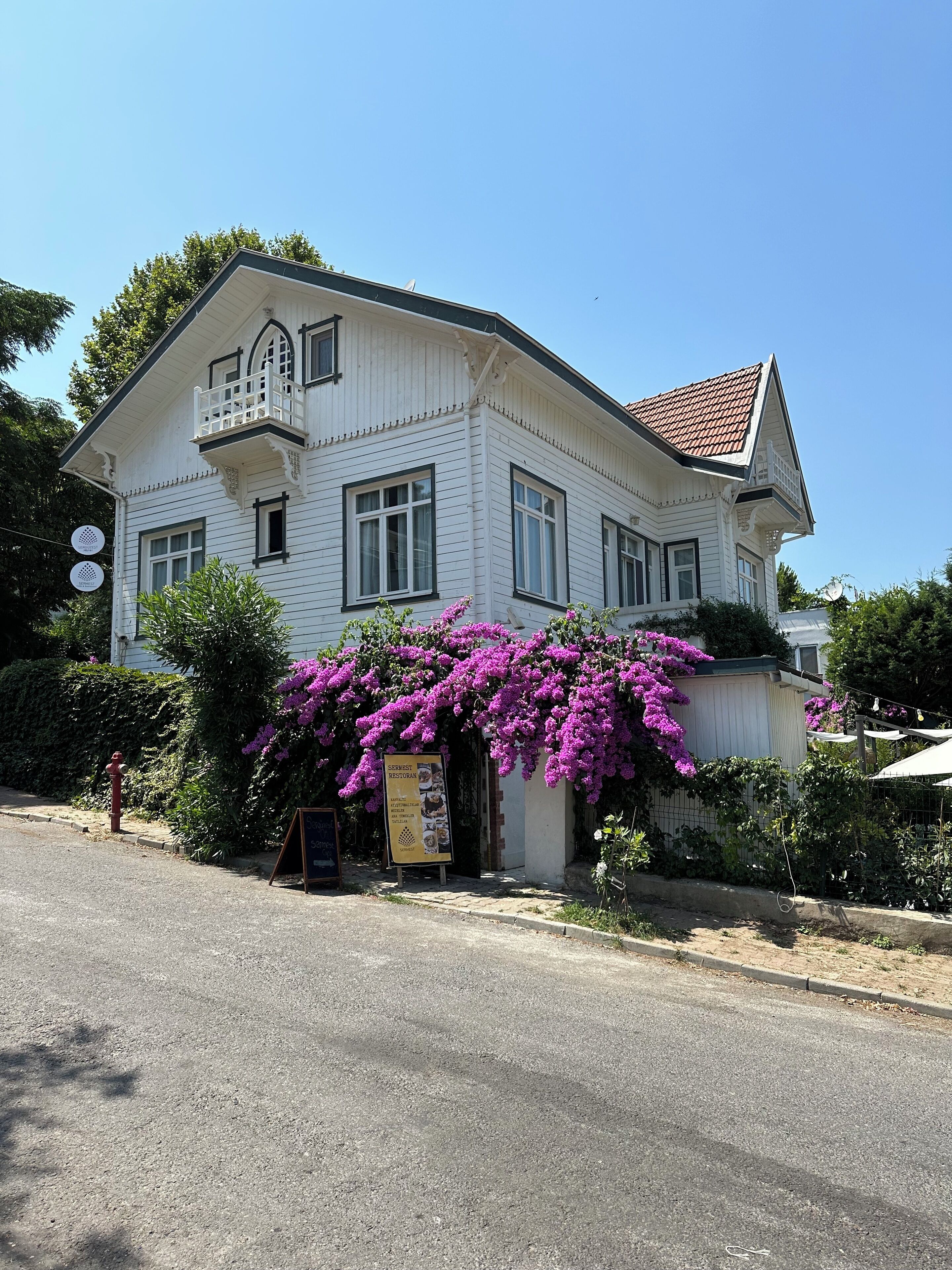 A white building with a balcony, a signboard, and purple flowering plants in front.