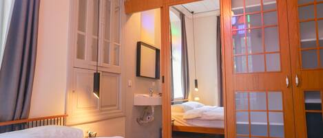 A hotel room with a bed, a wooden wardrobe, a mirror, a sink, and a window with curtains.