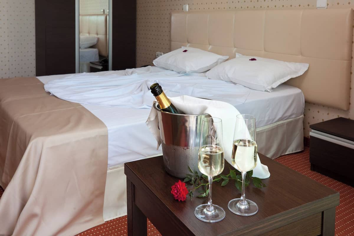 A hotel room with a bed, a bedside table with a champagne bottle, two wine glasses, and a rose.