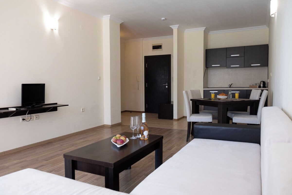 Una habitación de hotel moderna con sofá, una mesa de comedor con sillas, una zona de cocina y un televisor montado en la pared.