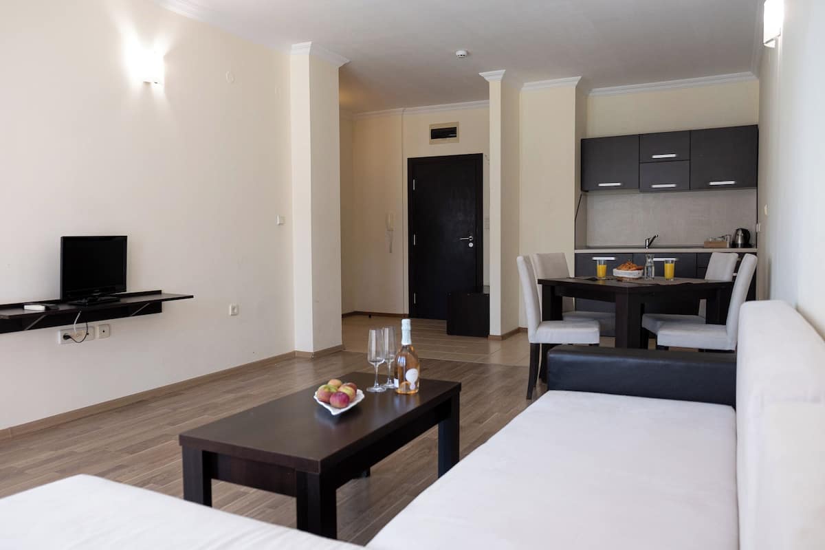 A modern hotel room with a sofa, a dining table with chairs, a kitchen area, and a TV mounted on the wall.