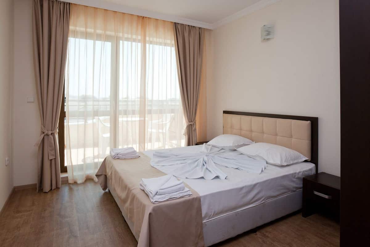 A hotel room with a bed, a nightstand, and a view of the beach through sheer curtains.
