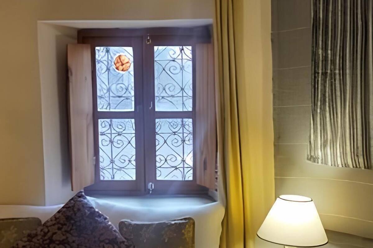 Ett hotellrum med en säng, ett fönster med dekorativa järngaller, en sänglampa och ett litet bord med en näsdukslåda.