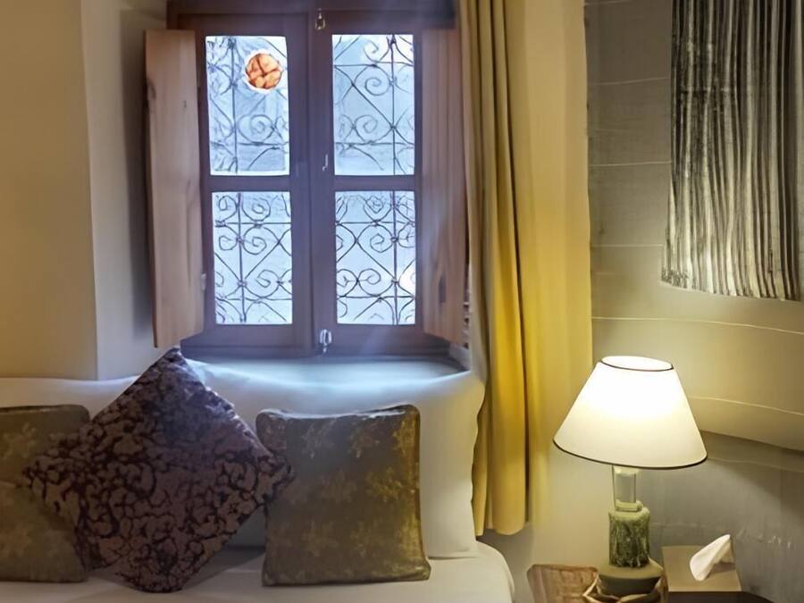 Una camera d'albergo con un letto, una finestra con inferriate decorative, una lampada da comodino e un piccolo tavolino con un pacchetto di fazzoletti.