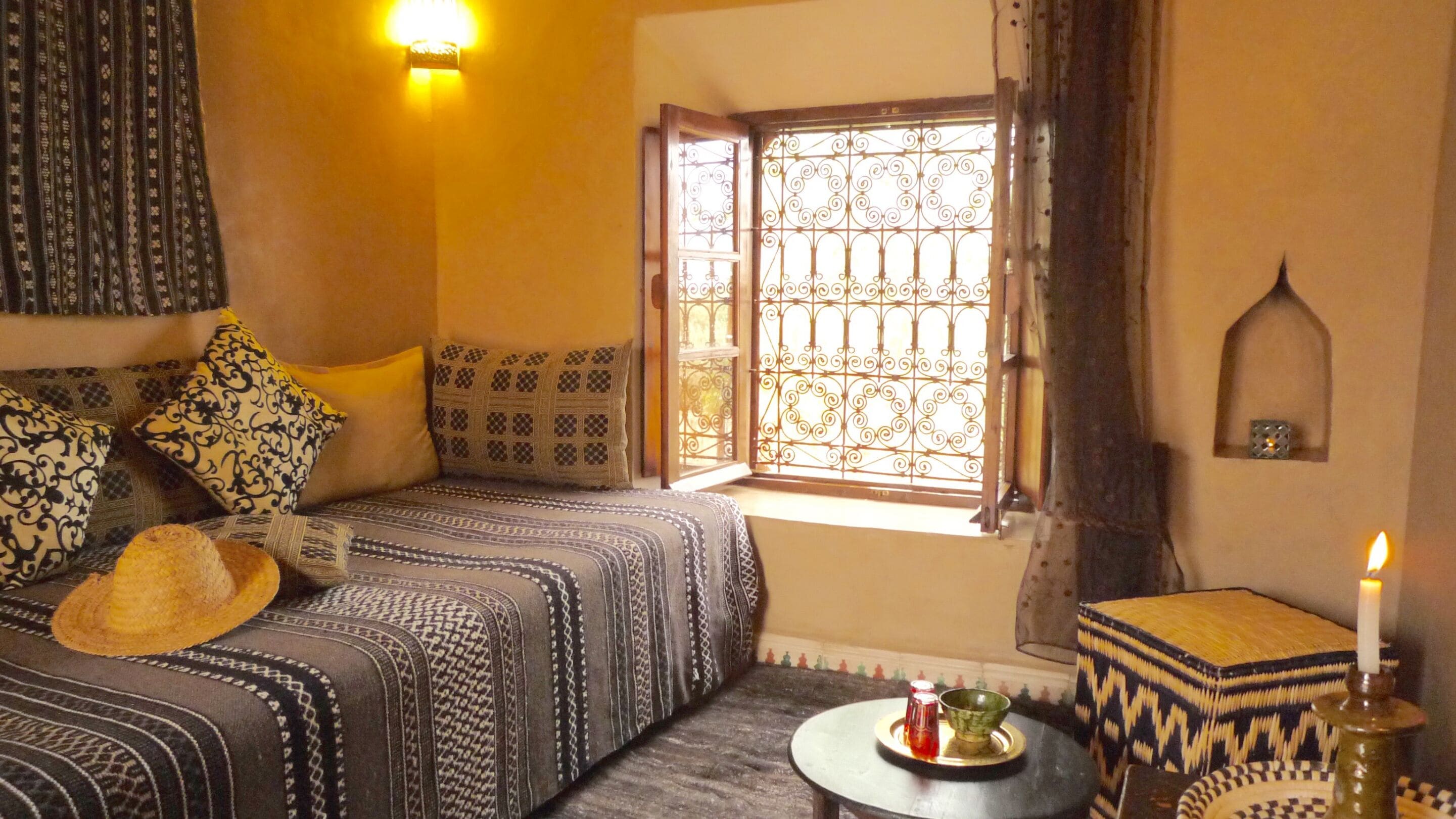 A cozy room with a bed, pillows, a small table with a candle, and a window with decorative iron bars.