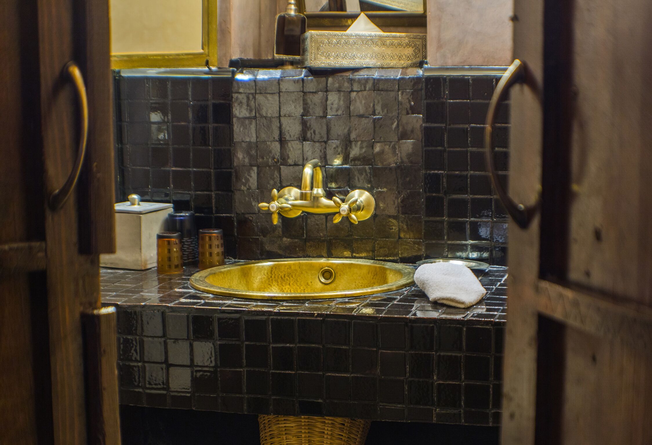 Un baño con pared de azulejos negros, un lavamanos dorado y una puerta de madera.
