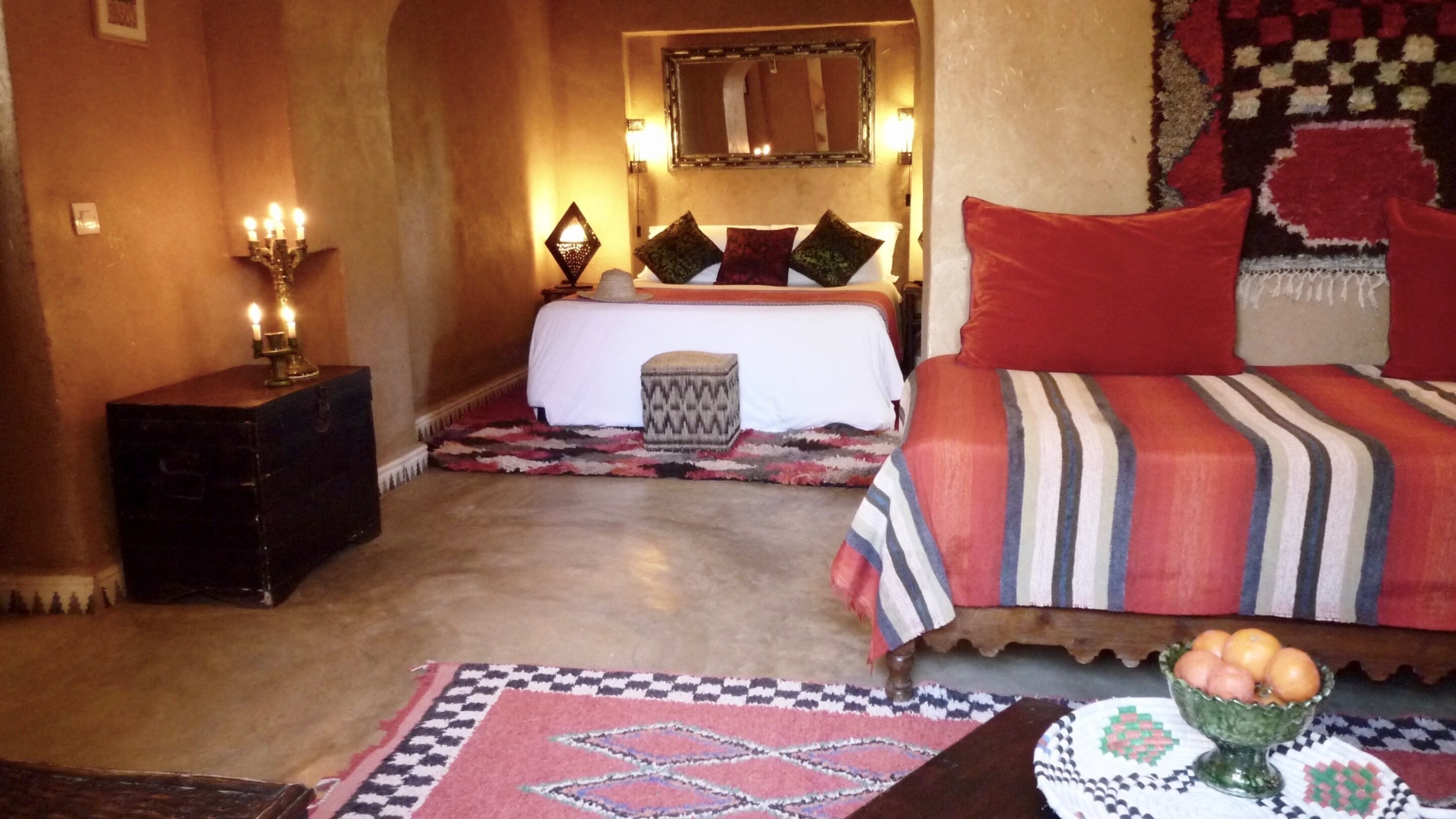 A room with a bed, a nightstand, a lamp, a mirror, and a small table with fruit.