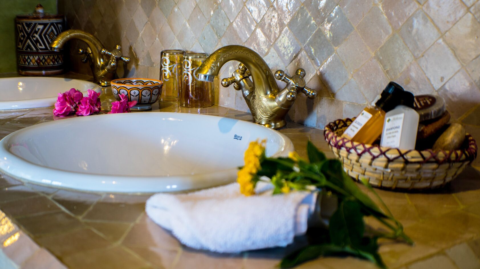 Un lavamanos con grifo dorado, un lavabo blanco y un cuenco decorativo con artículos de aseo.
