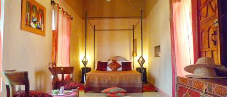 A four-poster bed with a canopy, a side table with a lamp, a chair, and a decorative hat on a stand.