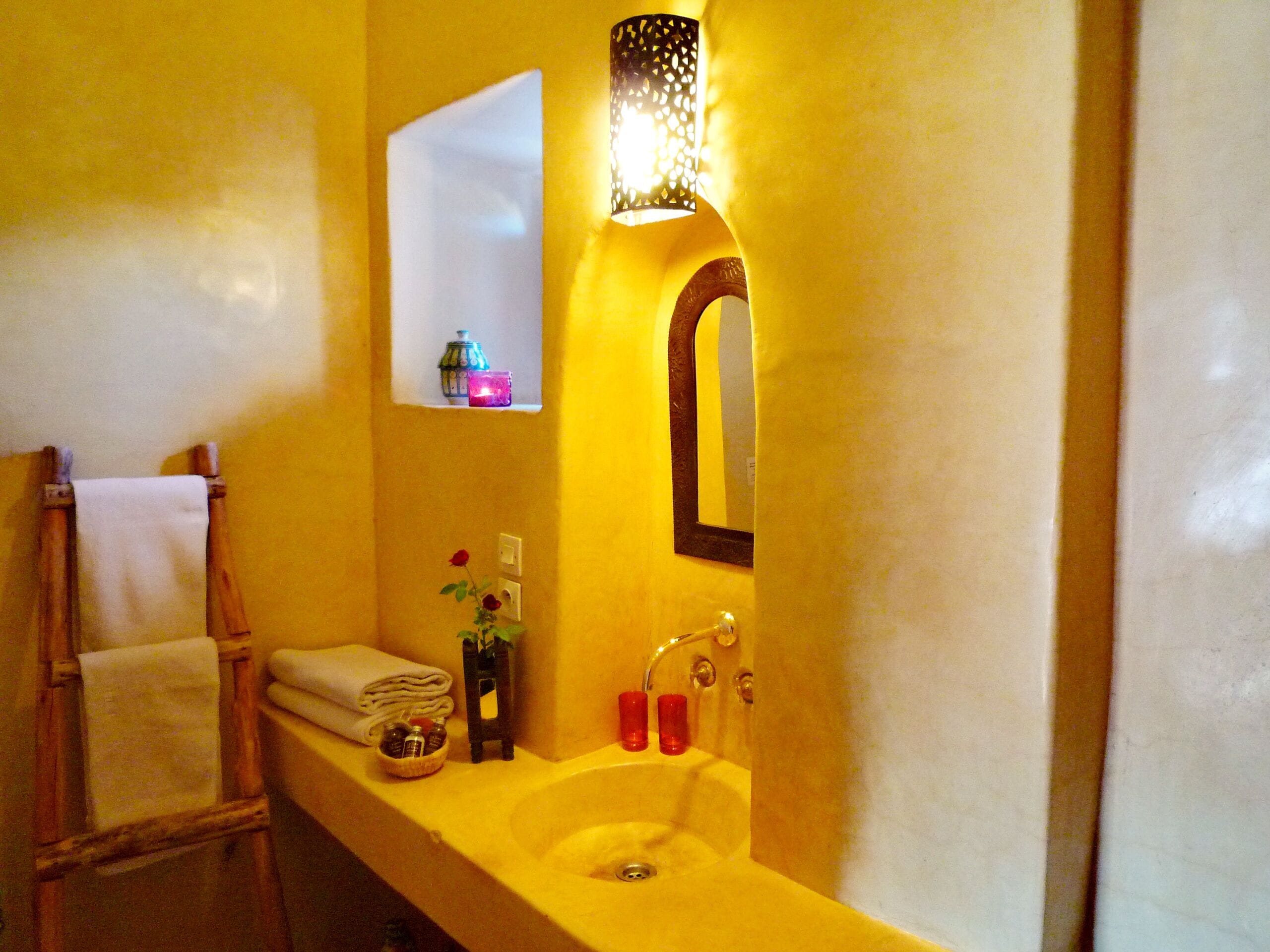 Un baño con un lavamanos amarillo, un espejo, un perchero y un estante pequeño con una vela y una botella.