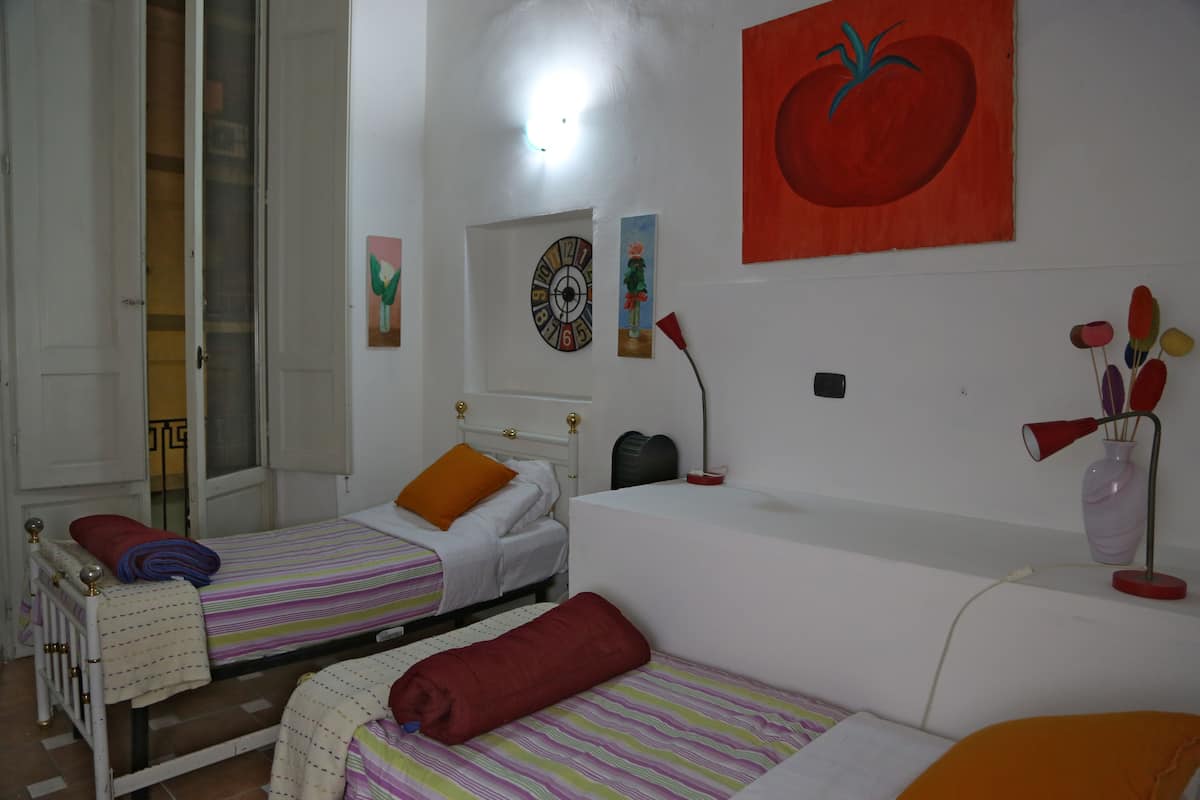 A bedroom with two single beds, a white sofa, a red tomato painting, and a wall clock.