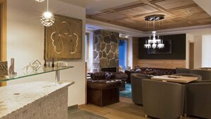 A reception area with a glass counter, a stone wall, and a stone fireplace. There are leather sofas and a chandelier in the background. - Chalet Marcora (Campitello di Fassa)