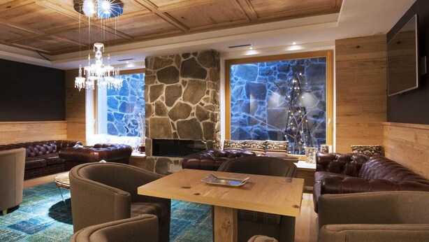 A cozy living room with a stone fireplace, leather sofas, a wooden dining table, and a chandelier. - Chalet Marcora (Campitello di Fassa)