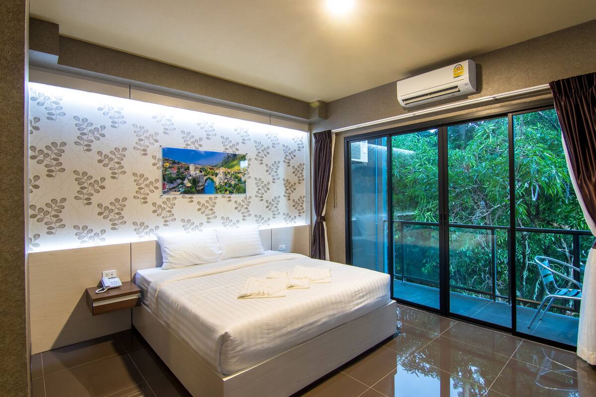 A modern hotel room with a large bed, a nightstand, a wall-mounted air conditioner, and a view of greenery through large windows.