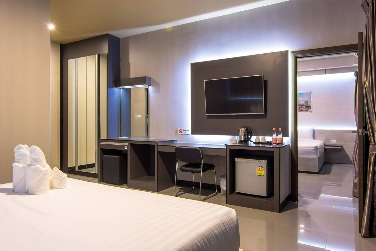 A modern hotel room with a large bed, a desk with a chair, a flat-screen TV, a minibar, and a small refrigerator.
