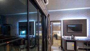 A modern hotel room with a glass partition, a flat-screen TV, a mini-fridge, a desk, and a seating area with a sofa and a small table. - PSG Hotel (Udon Thani)