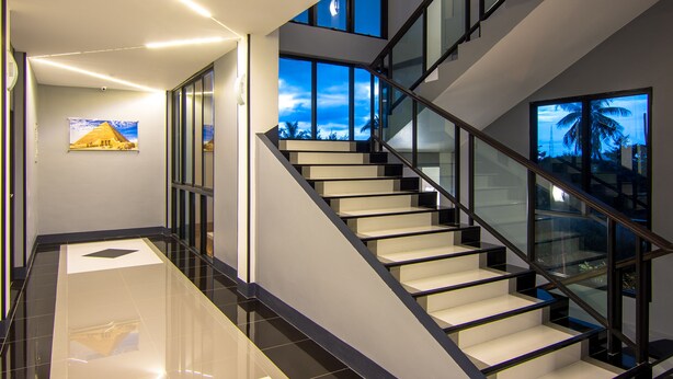 A modern staircase with glass railings and a view of palm trees through the windows. - PSG Hotel (Udon Thani)