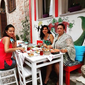 Trois personnes sont assises à une table, devant divers plats et boissons. On aperçoit un mur de briques et une fresque murale en arrière-plan.
